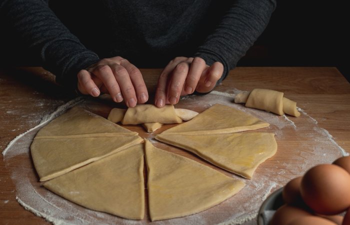 Close-up of hands rolling dough into croissants on a floured surface.