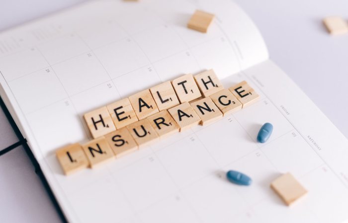 Close-up of health insurance spelled out with Scrabble tiles on a planner, alongside medication pills.