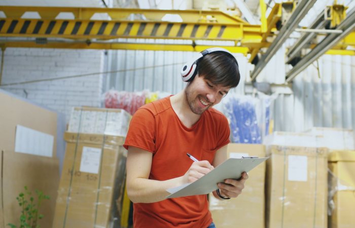 Smiling warehouse worker wearing headphones and writing on clipboard