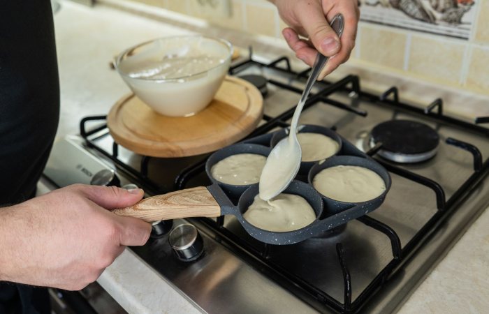 Close-up of a person preparing pancakes on a stovetop using a ladle and batter-filled skillet.