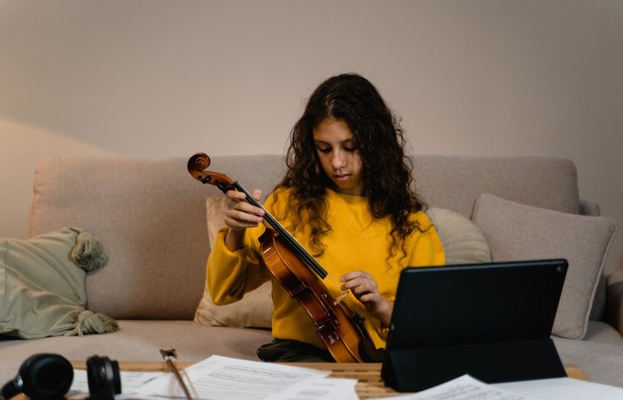 Teen girl in yellow sweater playing violin on couch with sheet music and tablet.