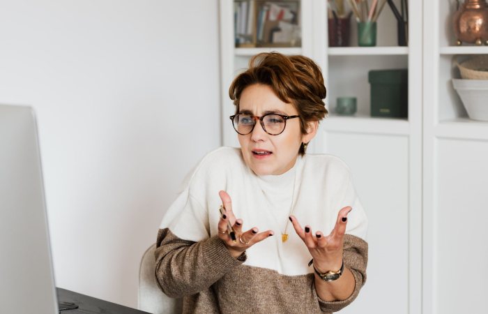 Expressive middle aged woman in casual outfit and eyeglasses gesticulating while having video conversation on computer in modern office