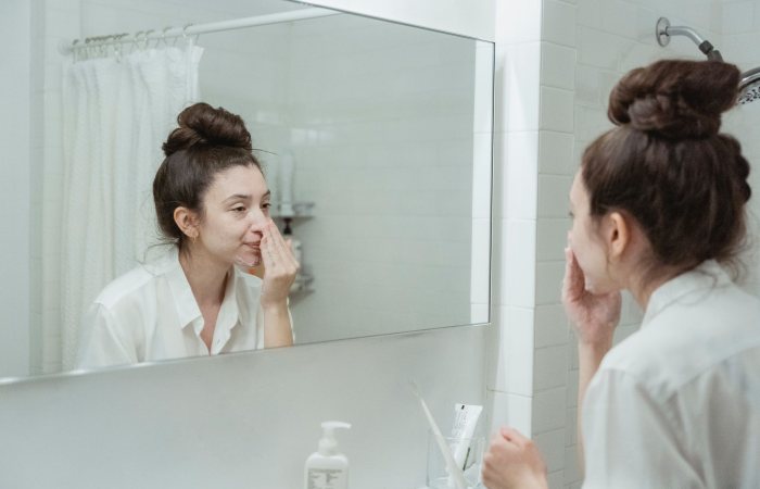 Caucasian woman in bathroom applying skincare product while looking in the mirror.